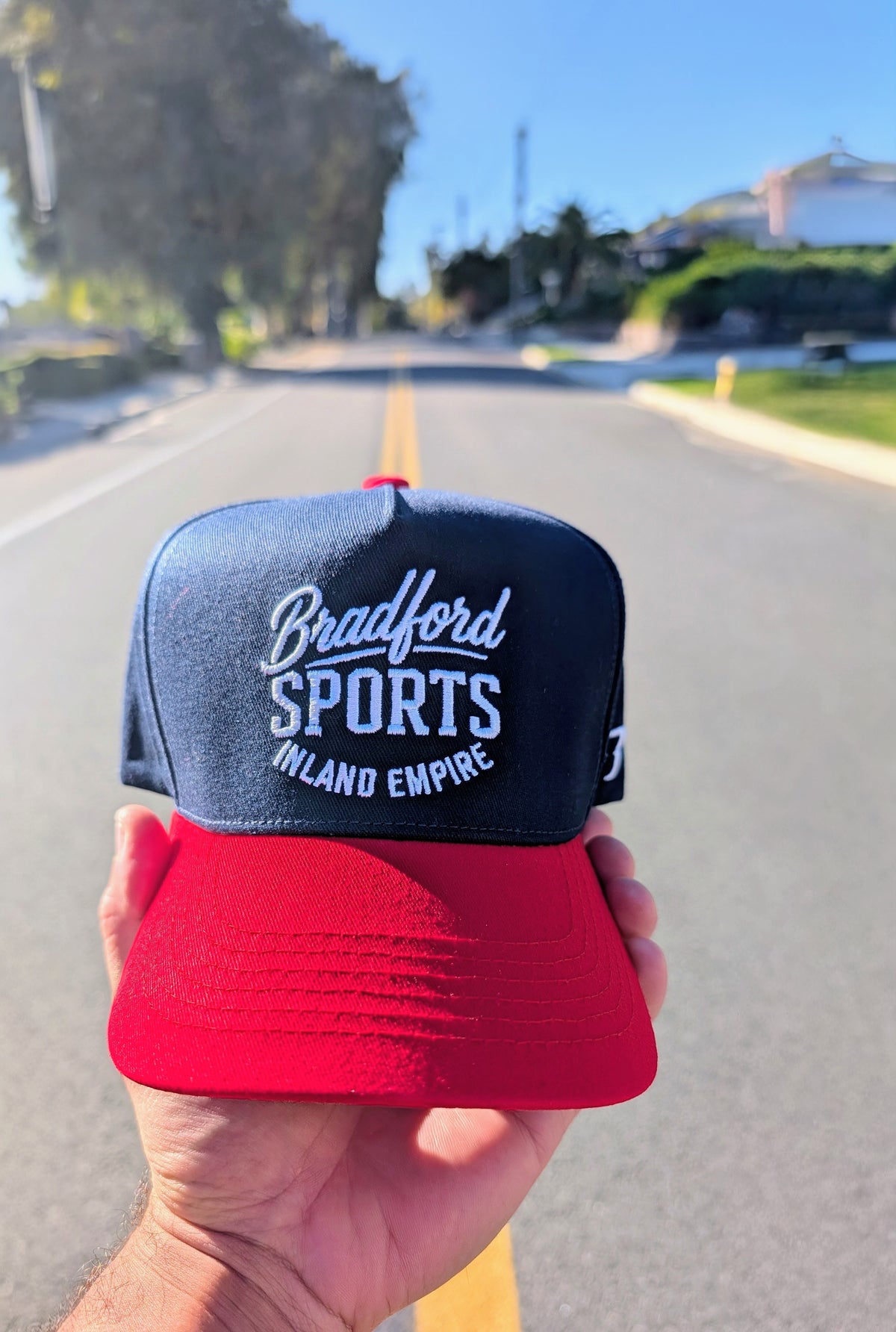 Hand holding the Bradford Sports Hometown trucker hat in red and navy, palms and an Inland Empire street through the windshield