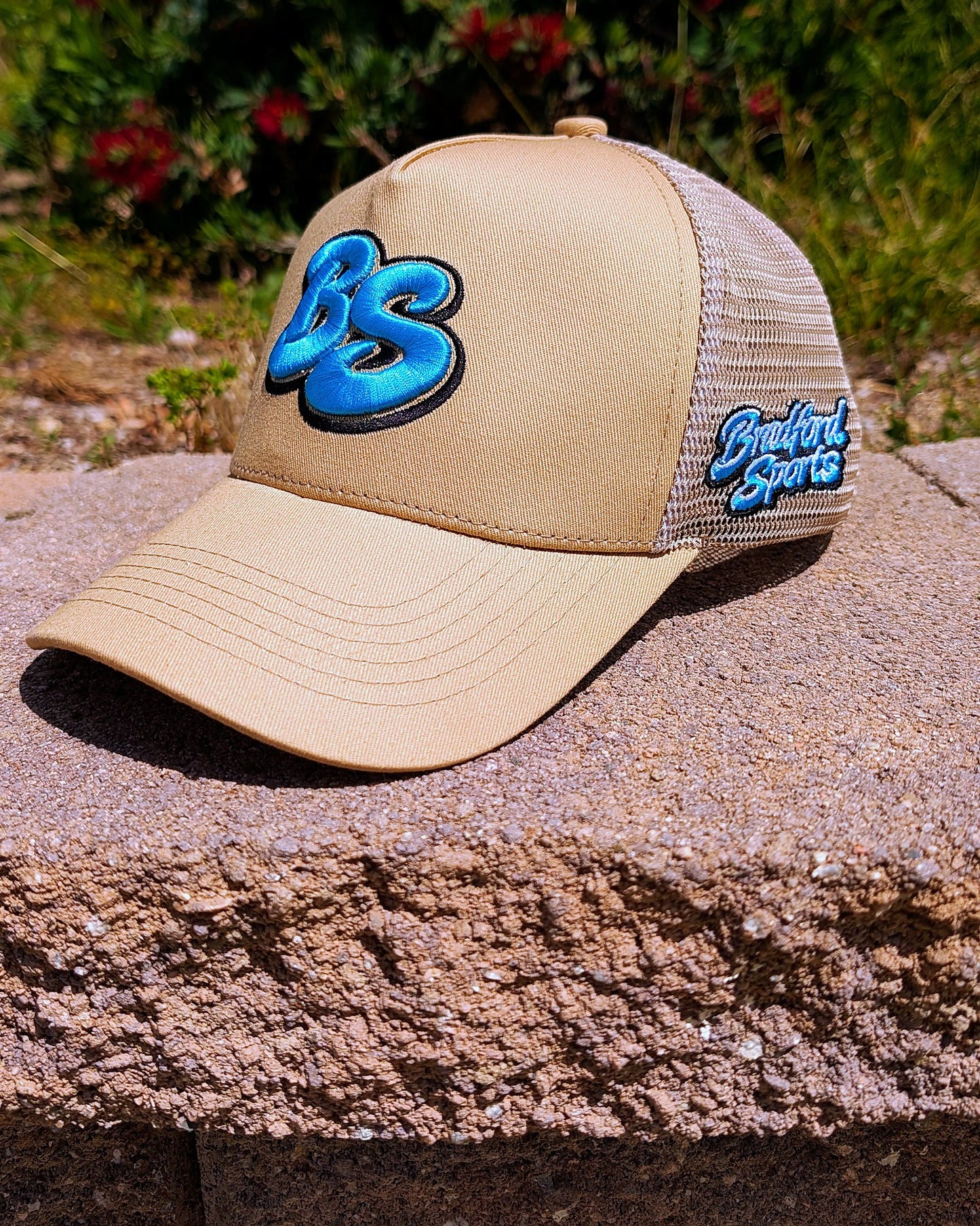 Bradford Sports Essential trucker hat in beige with teal BS puff embroidery on the front panel and tan mesh back, on a concrete wall in Norco, California