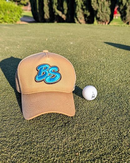 Beige Essential trucker hat resting on a putting green beside a golf ball in late afternoon light